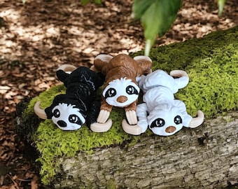 Puede incluir: Tres figuras de perezosos en negro, marrón y blanco con garras y rasgos faciales color canela descansan sobre un tronco cubierto de musgo. Los perezosos tienen ojos grandes y expresivos y están tumbados. El fondo incluye hojas y un entorno natural borroso.