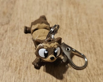 May include: A brown and tan raccoon-shaped keychain featuring large white eyes with black pupils, a small black nose, and pointed ears. The figure is attached to a silver-toned metal split ring and a lobster claw clasp for secure attachment.
