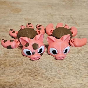 May include: A collection of pink and brown 3D-printed animal toys, including pig heads with dark brown spots and large eyes, and turtle-like bodies with matching pink and brown details. The toys are arranged on a wooden surface.