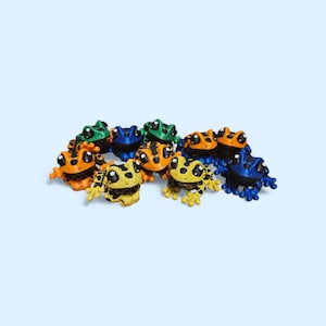 Puede incluir: Una colección de pequeñas figuras de ranas articuladas en varios colores, incluyendo naranja, amarillo, azul y verde. Cada rana presenta ojos grandes y redondos, un patrón de manchas en la espalda, extremidades articuladas y una cabeza ancha.