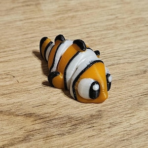 May include: A 3D-printed clownfish figurine. The fish features orange, white, and black stripes and markings. The figurine is lying on its side, with its large, round eyes visible. The fish is a decorative item.