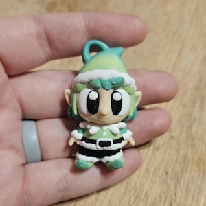 May include: A small, 3D-printed elf figurine with a green and white color scheme. The elf has large, expressive eyes, a pointed hat, and a belt. The figurine is held in a hand.