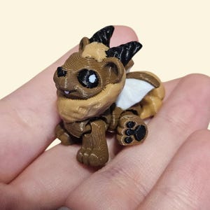 May include: A 3D-printed figurine of a fantasy creature, featuring a brown body with darker brown wood-grain texture on the legs and chest. The creature has black horns, white accents, and large eyes with white highlights. The figurine is held in a hand.