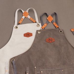 Può includere: Due grembiuli con spalline regolabili e dettagli in pelle. Uno è bianco sporco con "DK" su una toppa in pelle, l'altro è marrone con "DELBERT". Entrambi hanno elementi in ottone.