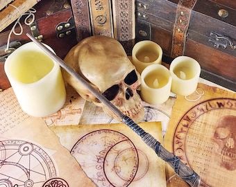 May include: A wooden box with a brass latch, a skull, a wand, and candles on a table with parchment paper featuring a circle and text. The text on the parchment reads "Book of Spells".