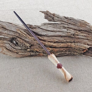May include: A dark brown wooden wand with a light brown handle and a red circular design. The wand is resting on a piece of driftwood.