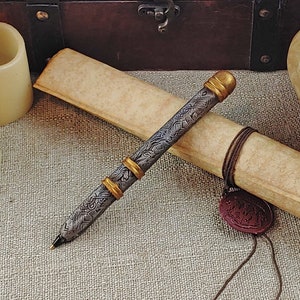 May include: A silver pen with gold accents and an intricate design. The pen is lying on a piece of parchment paper with a brown leather cord and a wax seal.