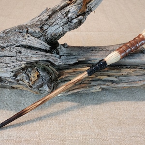 May include: A wooden wand with a brown leather wrap and a white bone tip. The wand is resting on a piece of driftwood.