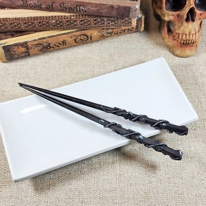 May include: Two black wands with intricate rose and vine detailing rest on a white rectangular plate. The wands are placed on a beige textured surface, with antique books and a skull in the background.