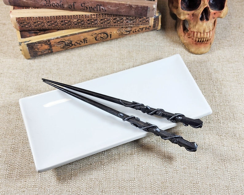 May include: Two black wands with intricate rose and vine detailing rest on a white rectangular plate. The wands are placed on a beige textured surface, with antique books and a skull in the background.