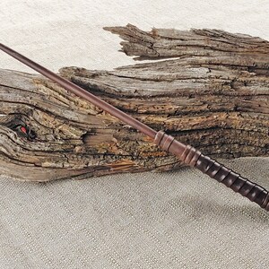 May include: A dark brown wooden wand with a textured grip. The wand is lying on a piece of driftwood.