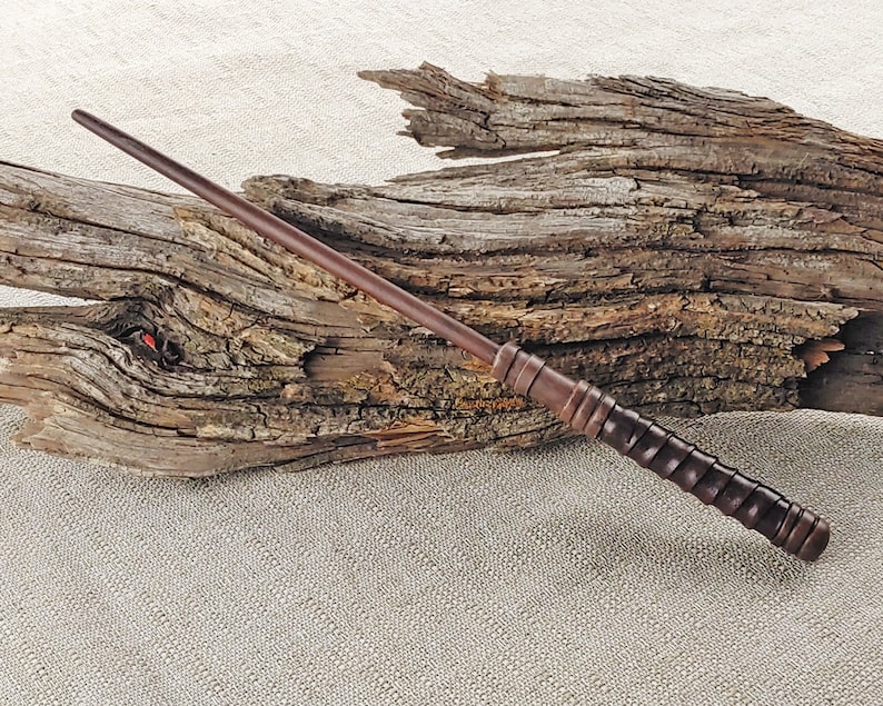 May include: A dark brown wooden wand with a textured grip. The wand is lying on a piece of driftwood.