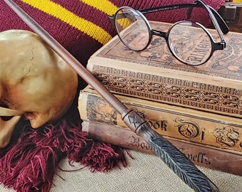 May include: A brown wooden wand with a dark feather detail, resting on top of a stack of old books with a skull and a pair of round glasses in the background.