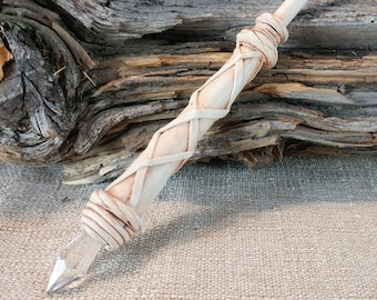 May include: A white wooden wand with a clear crystal tip and a woven leather design. The wand is resting on a piece of driftwood.