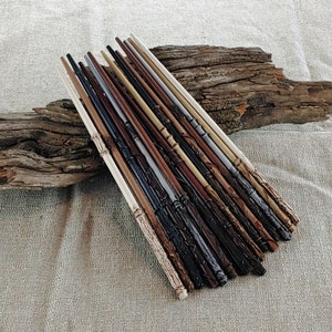 May include: A collection of handcrafted wooden wands in various colors, including brown, black, and cream. Each wand features unique carved details and textures. The wands are arranged on a piece of driftwood, set against a neutral fabric background.