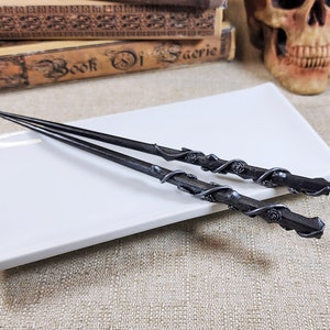 May include: Two dark gray wands with intricate floral and vine detailing, resting on a white rectangular plate. The wands have a textured appearance and are likely made of metal or a similar material. The overall theme suggests a fantasy or magical aesthetic.
