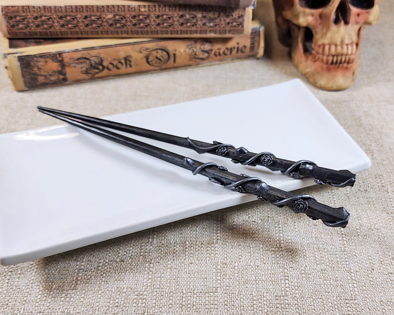 May include: Two dark gray wands with intricate floral and vine detailing, resting on a white rectangular plate. The wands have a textured appearance and are likely made of metal or a similar material. The overall theme suggests a fantasy or magical aesthetic.