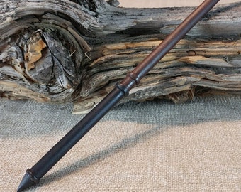 Puede incluir: Una varita de madera marrón oscuro con una punta negra y una banda plateada cerca de la punta. La varita está apoyada en un trozo de madera a la deriva.