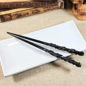 May include: Two black chopsticks with ornate, swirling silver-colored designs and rose accents rest on a white rectangular plate. The chopsticks are angled, with their tips pointing away from the viewer. The background includes antique books and a skull.