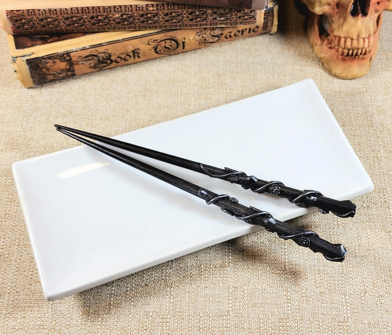 May include: Two black chopsticks with ornate, swirling silver-colored designs and rose accents rest on a white rectangular plate. The chopsticks are angled, with their tips pointing away from the viewer. The background includes antique books and a skull.