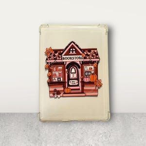 Puede incluir: Una funda de tableta de color crema con una ilustración caprichosa de una librería. La librería tiene un techo marrón, paredes rosas y está decorada con calabazas, hojas y setas. La palabra "BOOKSTORE" se muestra encima de la entrada.