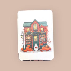 Puede incluir: Una tarjeta blanca con una colorida ilustración de un edificio de ladrillo con techo verde, calabazas y hojas de otoño. Una guirnalda de luces cuelga sobre la puerta. Un fantasma lee un libro frente al edificio.