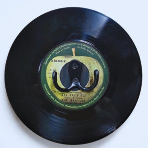 Peut inclure: Un disque vinyle noir avec un crochet en métal noir. Le disque présente une étiquette verte en forme de pomme avec du texte, dont "GET BACK" et "THE BEATLES". Le crochet est conçu pour suspendre des objets.