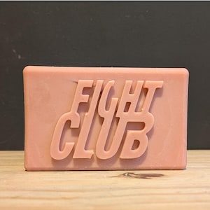 Könnte beinhalten: Ein rechteckiger Block rosa Seife mit dem geprägten Schriftzug "FIGHT CLUB" auf der Oberfläche in einer fetten, serifenlosen Schriftart. Die Seife hat eine einfarbige Farbe und scheint unparfümiert zu sein. Die Seife liegt auf einer Holzoberfläche.