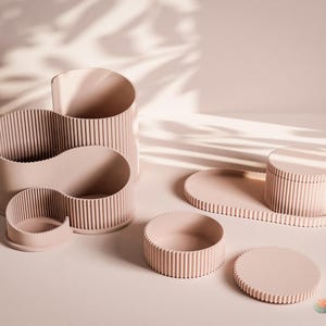 Könnte beinhalten: Ein Set aus hellrosa Schreibtisch-Organizern und Aufbewahrungsbehältern mit vertikaler Rippenstruktur. Die Kollektion umfasst einen Stiftehalter mit mehreren Fächern, eine kleine runde Schale, einen Behälter mit Deckel auf einem ovalen Tablett.