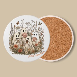 Pode incluir: Um porta-copos redondo com um topo branco apresentando quatro fantasmas brancos num campo de flores silvestres e borboletas. Os fantasmas seguram uma chávena de café, uma concha rosa e uma caneca castanha. O texto pan-tasmas está em laranja.