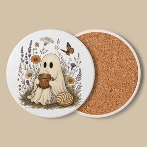 Puede incluir: Un posavasos redondo con un fantasma blanco sosteniendo una taza marrón con vapor, rodeado de flores silvestres coloridas y una mariposa. Junto al fantasma hay un pan redondo texturizado. El posavasos tiene una base de corcho natural.