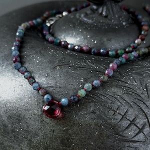 Könnte beinhalten: Halskette mit bunten Edelsteinen. Die Kette hat eine rote Schnur und einen großen, facettierten, tropfenförmigen Anhänger. Die Edelsteine sind in Blau-, Grün- und Rottönen gehalten. Ein Schmuckstück.