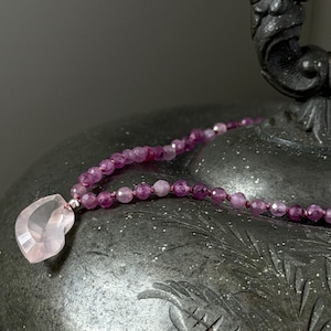 May include: A delicate necklace featuring a faceted, heart-shaped rose quartz pendant. The necklace is strung with small, round, purple beads. The jewelry is displayed on a dark, textured surface, creating a contrast with the light pink and purple hues.