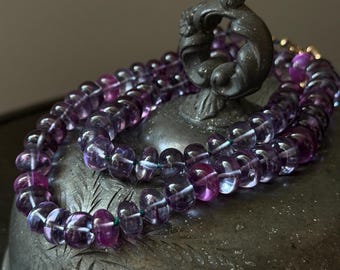 May include: A necklace of translucent purple beads with a dark metal clasp. The beads are strung on a green thread and rest on a dark, textured surface. The necklace is draped over a decorative metal object.