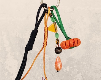 May include: A decorative keychain featuring a pumpkin design. The keychain includes a silver ring, black and orange cords, and a small orange pumpkin charm. Additional charms include a yellow triangle, green beads, and a teardrop-shaped stone. The text "Pumpkin 01" is visible.