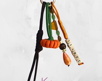 May include: A keychain with a gold ring, featuring a small orange pumpkin charm, green and orange cords, and wooden beads spelling "ISABEL". The keychain also includes a brown and orange teardrop-shaped bead. The text "Pumpkin 03" and "Keytique" are visible.