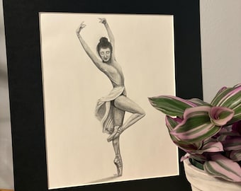 Puede incluir: Dibujo a lápiz de una bailarina en una pose elegante, enmarcado en un passe-partout negro. La bailarina lleva un maillot y una falda fluida, con los brazos extendidos por encima de la cabeza. La obra de arte se exhibe cerca de una planta en maceta.