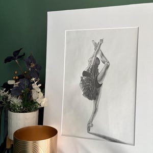 Puede incluir: Un dibujo a lápiz enmarcado de una bailarina en una pose dramática, con una pierna extendida hacia arriba. La bailarina lleva un tutú. La obra de arte está sobre un paspartú blanco y una pared verde oscuro. Un recipiente decorativo dorado y una planta en maceta están en primer plano.