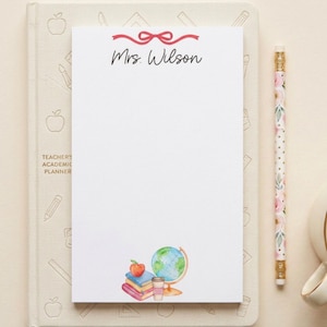 May include: A white notepad with the text "Mrs. Wilson" and a red bow design. The notepad features a watercolour illustration of a globe, books, an apple, and a coffee cup. A floral pencil and a coffee cup are also visible.