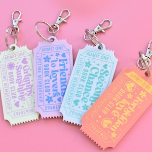 May include: Four colorful ticket-shaped keychains with silver clasps. Each keychain has a different pastel color and text: "Grumpy Sunshine", "Friends to Lovers", "Second Chance", and "Forbidden Love". The keychains are for a book club.