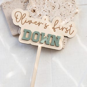 May include: A wooden cake topper with the text "Oliver's first DOWN" in a combination of script and block letters. The word "DOWN" is in a muted green color. The topper has a long, thin wooden stick for insertion into a cake.