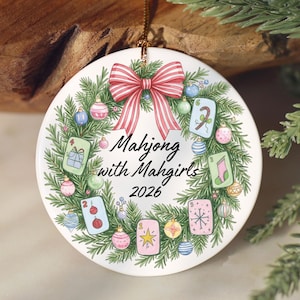 Puede incluir: Un adorno redondo de cer&aacute;mica blanca con una corona de pino verde decorada con bolas de Navidad coloridas y fichas de Mahjong. Un lazo a rayas rojas y blancas corona la parte superior. El texto dice Mahjong with Mahgirls 2026 en negro.