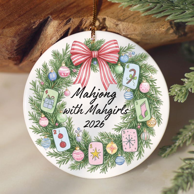 Puede incluir: Un adorno redondo de cer&aacute;mica blanca con una corona de pino verde decorada con bolas de Navidad coloridas y fichas de Mahjong. Un lazo a rayas rojas y blancas corona la parte superior. El texto dice Mahjong with Mahgirls 2026 en negro.