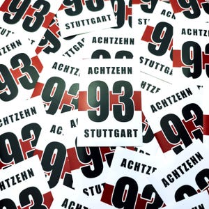 Op de afbeelding: Een verzameling witte rechthoekige stickers met het zwarte nummer "93", een rode streep en het zwarte woord "STUTTGART". Sommige stickers hebben ook het woord "ACHTZEHN".