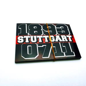 Op de afbeelding: Een stapel zwarte en rode stickers met de tekst "1893 STUTTGART 0711" in het wit. De stickers worden bij elkaar gehouden door een gele elastiek. De achtergrond is wit.