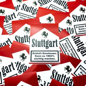 Op de afbeelding: Een verzameling rode en witte rechthoekige kaarten met het woord "Stuttgart" in zwarte letters. Elke kaart heeft een zwart paardlogo en Duitse tekst, waaronder "Enthält Emotionen. Kann zu 1893% süchtig machen."