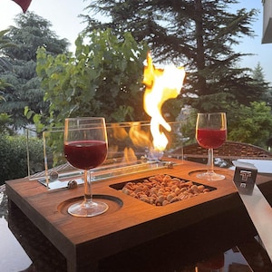 Puede incluir: Un brasero de mesa de madera con dos copas de vino tinto. El brasero tiene un cerramiento de vidrio y una sección llena de pequeñas piedras de color marrón claro. El fondo muestra árboles verdes.