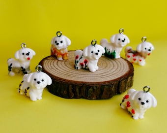 Puede incluir: Una colección de pequeñas figuras de perros blancos con ojos y narices negras, cada una adornada con un lazo plateado para colgar. Algunos perros presentan diseños florales o estampados coloridos en sus cuerpos, sobre un fondo amarillo.