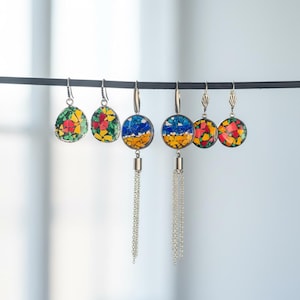 Puede incluir: Una colección de pendientes de mosaico coloridos. Algunos pendientes son redondos con cadenas plateadas, mientras que otros son triangulares. Los diseños de mosaico presentan colores rojo, amarillo, verde y azul. Los pendientes se exhiben en una barra negra.