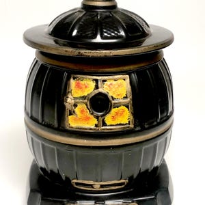 May include: A black ceramic cookie jar shaped like a vintage potbelly stove. The jar features a removable lid, a square window with yellow and orange painted flames, and metallic bronze-colored trim along the edges and base of the structure.